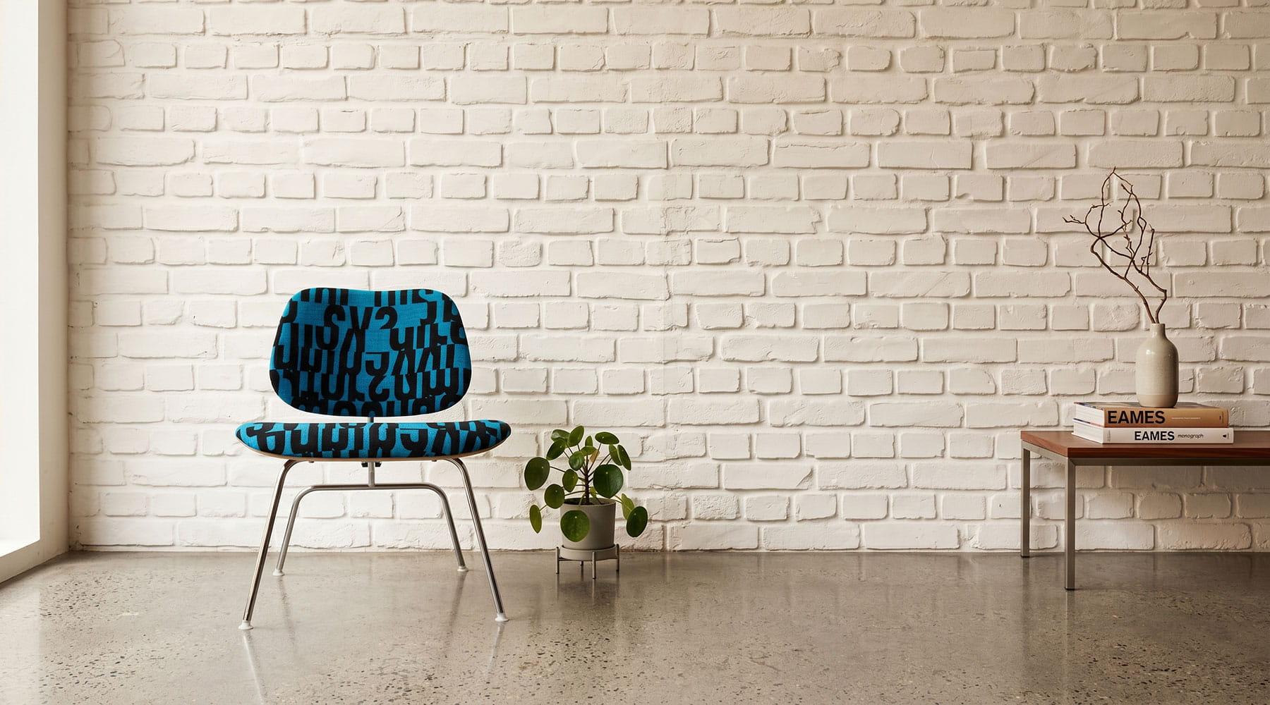 A blue patterned Eames Upholstered LCM chair against a white brick wall and concrete floor.