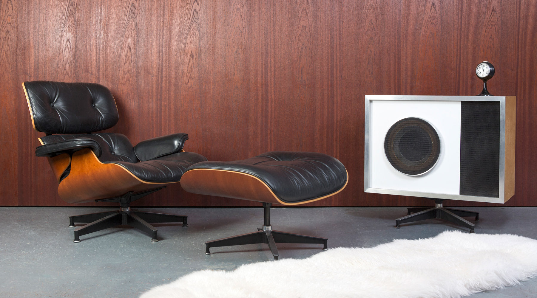 A gorgeous and original Eames Lounge Chair & Ottoman sit with a Trusonic Speaker against a wood paneled background